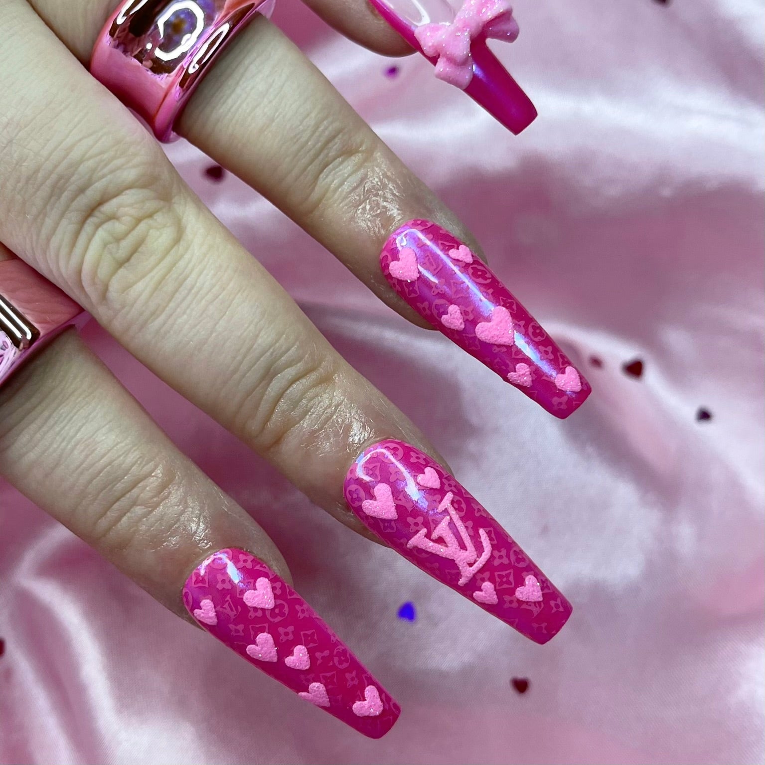 Louis Valentine press-on nails feature dark pink chrome French tips, glittered pink bows on the thumbs and pinkies, and embossed glittered logos and hearts for a romantic, luxury look.